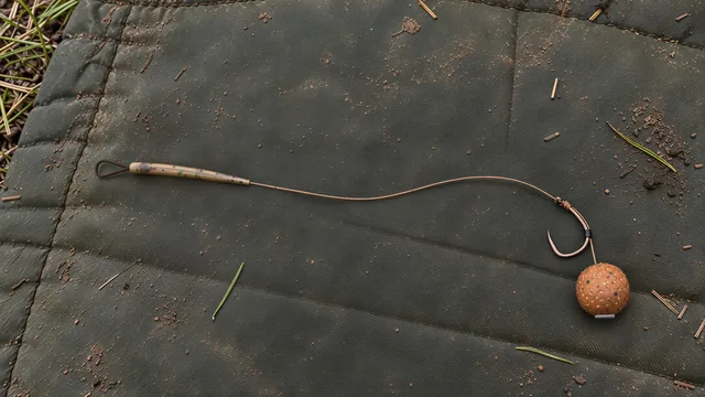 A hair rig lying on a worn green unhooking mat: a figure-of-eight loop under tapered camo shrink tube, about 15 cm of braid hooklink knotted to the eye of the hook, the line held along the back of the shank by a tiny silicone sleeve, and a boilie on the hair just below the bend with a flat bait stop across its base.